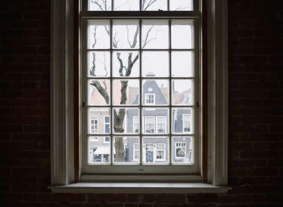 Een hoog oud houten schuifraam met kleine ruitjes in een dikke bakstenen muur, met uitzicht op een grachtenpand aan de overkant