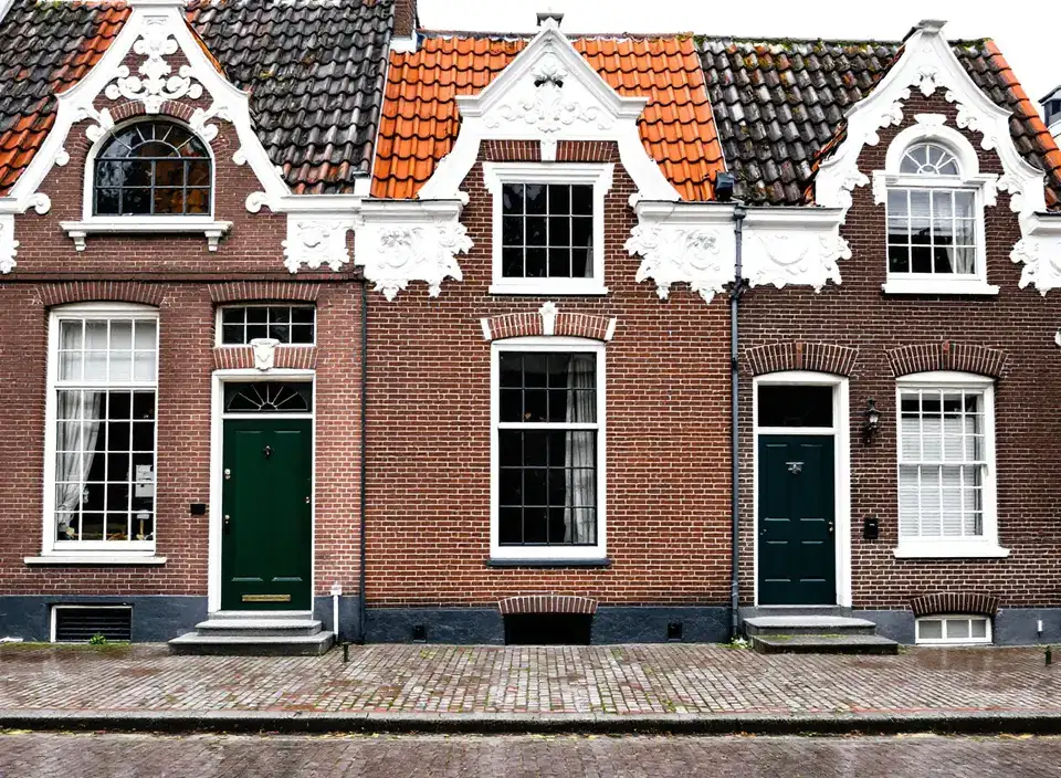 Drie oude bakstenen huizen naast elkaar met witte siergevels, hoge witte schuiframen en donkergroene voordeuren aan een straat met kinderkopjes