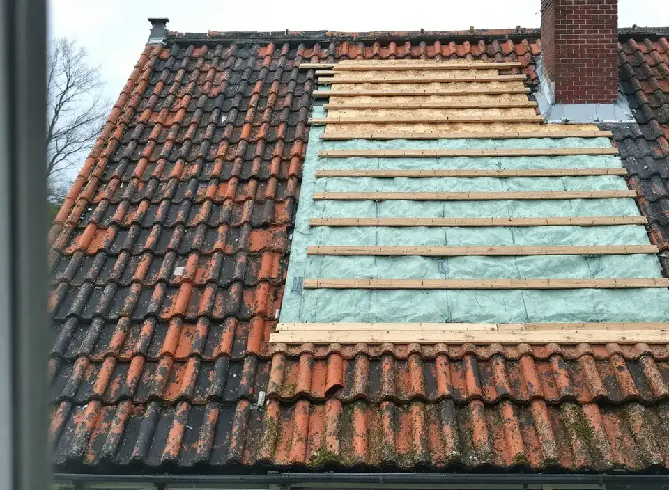 Een oud pannendak tijdens het werk: links de oude oranje pannen, rechts isolatieplaten onder nieuwe houten panlatten, met een bakstenen schoorsteen
