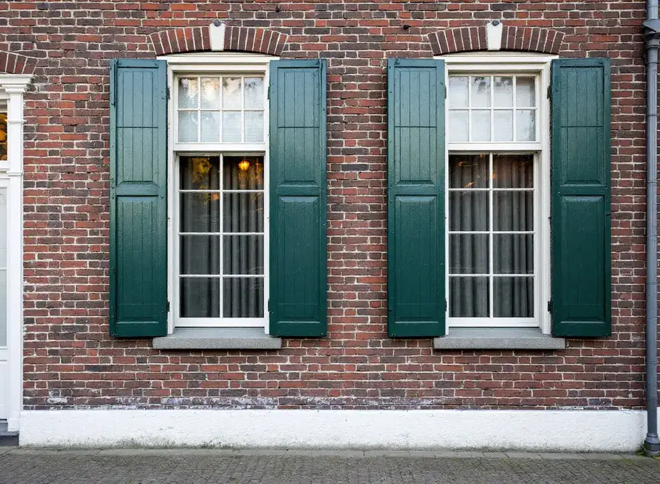 Twee hoge ramen in een bakstenen gevel met opengeslagen donkergroene houten luiken en een witte plint boven kinderkopjes